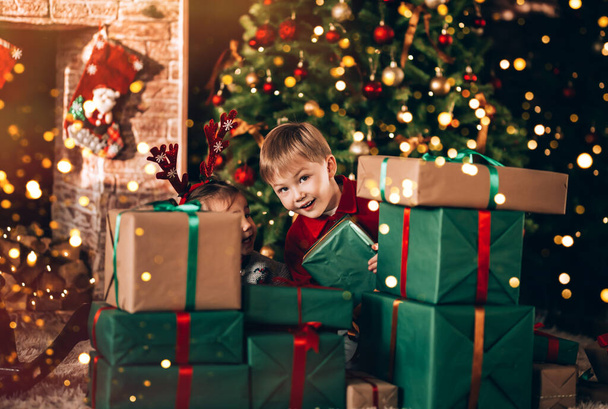 Brother and sister are sitting with boxes in green packaging with gifts and a bow. Christmas mood. Children in New Year's costumes waiting for a miracle. Against the background of a Christmas tree and fireplace. - Photo, Image