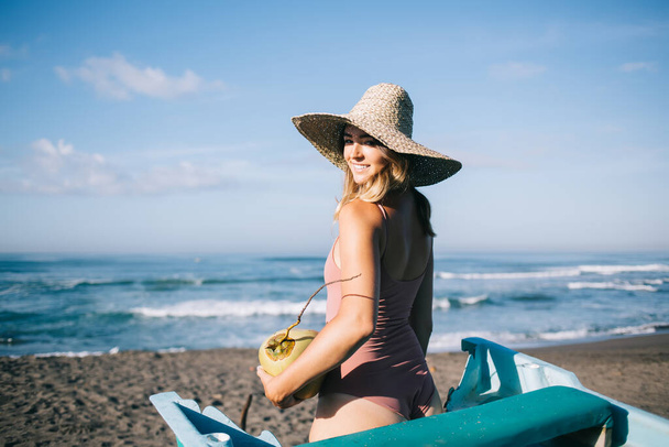 Side view of smiling female in hat and swimsuit standing on sandy beach with coconut near waving sea during sunny summer day and looking over shoulder - Foto, immagini