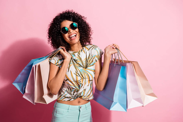 Portrait photo of happy smiling black skinned curly woman holding bags on black friday sunglass isolated on pastel pink color background - Zdjęcie, obraz