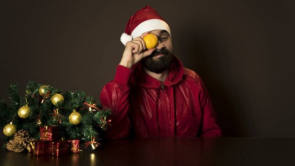 Ο νεαρός σύγχρονος Άγιος Βασίλης κάθεται σε ένα τραπέζι κοντά στο δέντρο. Συναισθηματικό πορτρέτο. Χριστούγεννα. Μανταρίνια - Φωτογραφία, εικόνα