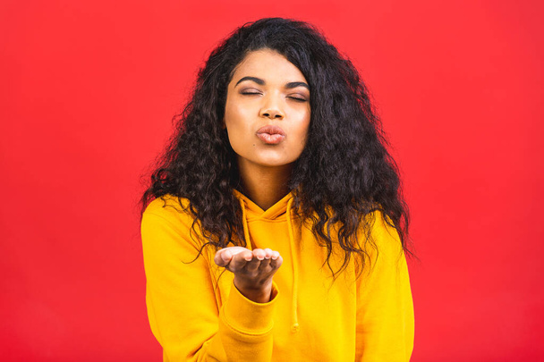 Pretty girl makes air kiss into camera, expresses her love. Photo of african american girl wears casual outfit on red background. Emotions and pleasant feelings concept. - Photo, Image