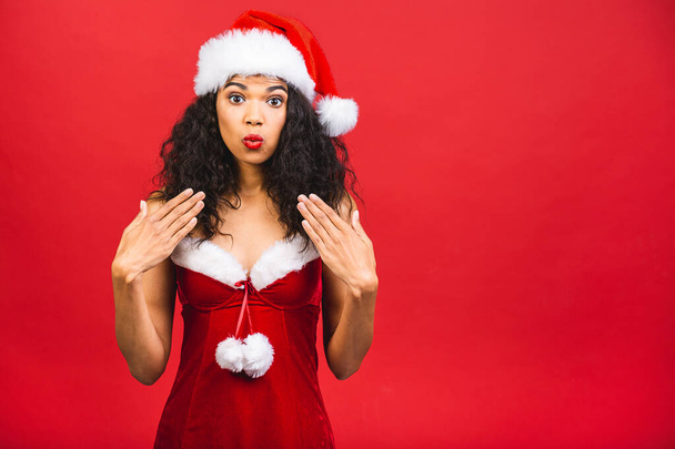 赤い背景の上に隔離されたサンタクロースの服で美しいショックを受けたアフリカ系アメリカ人の黒人女性。新年とクリスマスのコンセプト.  - 写真・画像