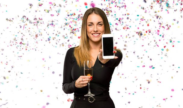 Женщина с шампанским празднует Новый год, смотрит в камеру и улыбается, пользуясь мобильным телефоном с конфетти - Фото, изображение