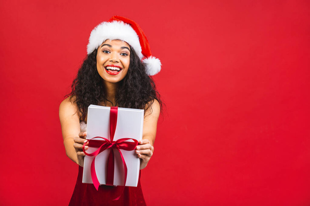 Gyönyörű mosolygós boldog afro-amerikai fekete nő Mikulás ruhák elszigetelt vörös háttér. Újév és karácsony koncepció. Ajándékdoboz.  - Fotó, kép