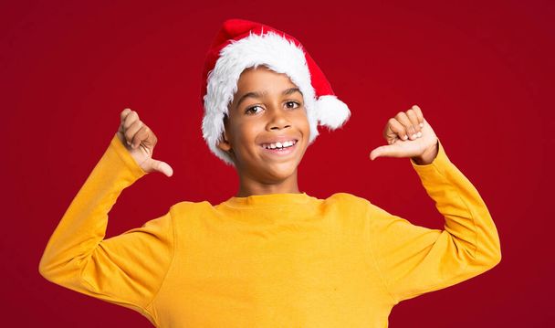 African American boy with christmas hat proud and self-satisfied over red background - Foto, imagen