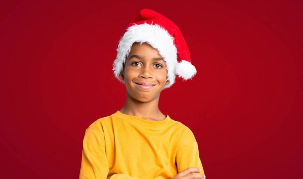 Menino afro-americano com chapéu de Natal sorrindo muito sobre fundo vermelho - Foto, Imagem