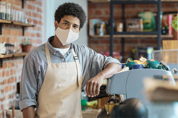 Young african american guy in apron and protective mask in modern loft coffee shop - Photo, Image