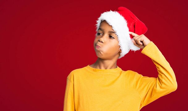 Menino afro-americano com chapéu de Natal com dúvidas e com expressão facial confusa sobre fundo vermelho - Foto, Imagem