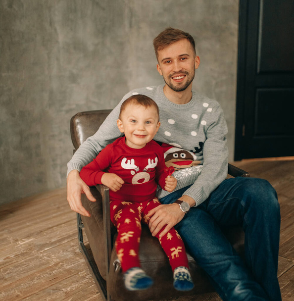 Father with his son sit in armchair in sweaters with Christmas ornament. - Фото, изображение