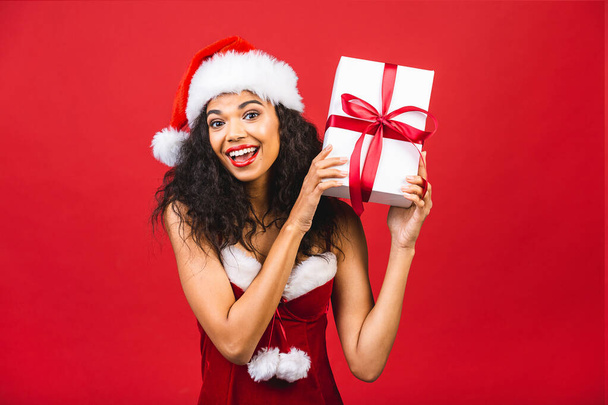 Gyönyörű mosolygós boldog afro-amerikai fekete nő Mikulás ruhák elszigetelt vörös háttér. Újév és karácsony koncepció. Ajándékdoboz.  - Fotó, kép