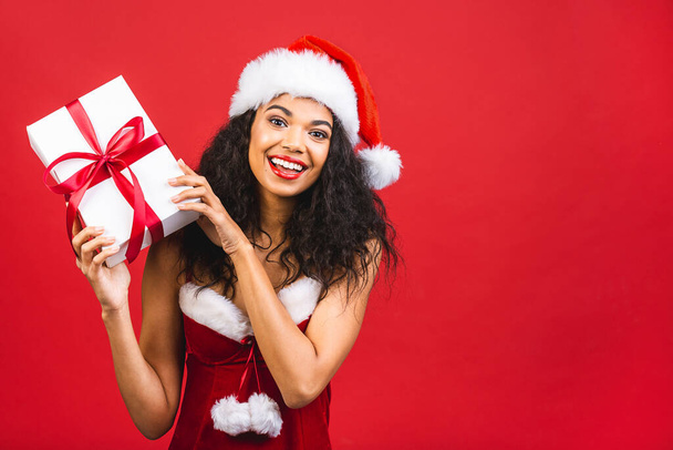 Гарна усміхнена щаслива афро-американська чорношкіра жінка в одязі Санта-Клауса ізольована на червоному тлі. Новий рік і християнська концепція. Тримаючи коробку з подарунками..  - Фото, зображення