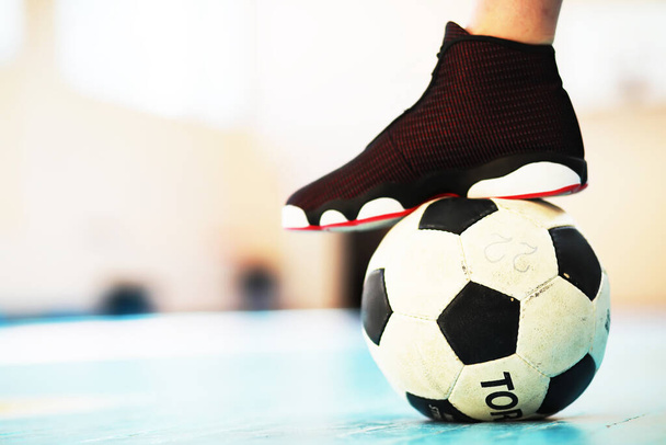 Un pie humano descansa sobre el balón en el suelo de hormigón. Foto de una pelota de fútbol y zapatillas en un piso de madera - Foto, Imagen
