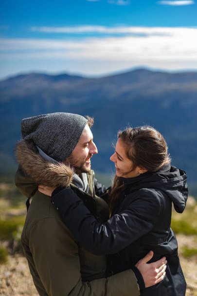 Aşık genç bir çift ve kışlık giysiler, güneşli bir günde dağ sırasının önünde sarılıp gülümsüyor. - Fotoğraf, Görsel