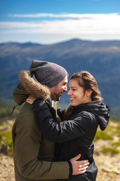 Young couple in love and in winter clothes hug and smile in front of the mountain range in a sunny day - Photo, Image