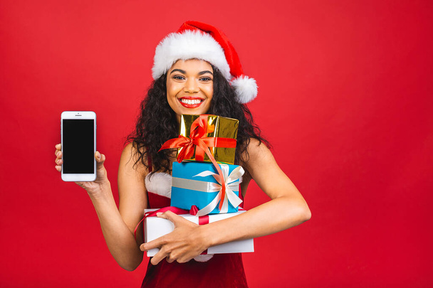 Glücklich aufgeregte Afroamerikanerin im roten Weihnachtsmann-Outfit mit Handy vor rotem Hintergrund. Geschenkboxen halten.  - Foto, Bild