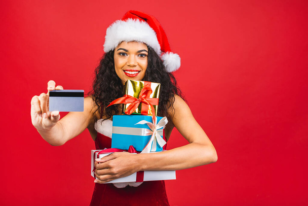 Beautiful portrait of young african american woman happy holding credit card and gift box in Christmas holiday isolated on red background, surprised girl shopping with credit card in xmas day. - Фото, зображення