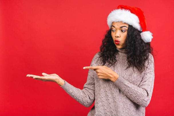 Portrait of beautiful african american female woman model wearing santa hat isolated over red background. Pointing finger.  - Photo, Image