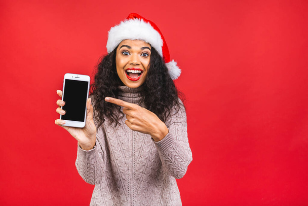 Felice donna afroamericana eccitato in rosso cappello di Babbo Natale con il telefono cellulare isolato su sfondo rosso. - Foto, immagini
