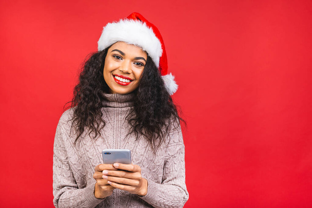 Happy excited african american woman in red santa claus hat with mobile phone isolated over red background. - Photo, Image