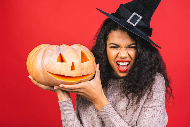 Portrait von Halloween afrikanisch-amerikanische Hexe Frau hält einen Kürbis und großen Hut isoliert über rotem Hintergrund. Halloween Stil, Halloween Party. - Foto, Bild