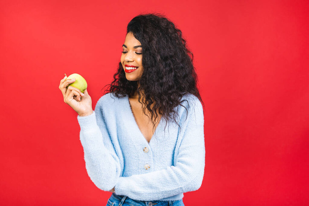 Jeune femme afro-américaine mangeant une pomme verte isolée sur fond rouge.  - Photo, image