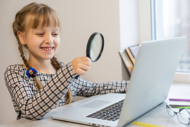 Evde, masada ödev yapan küçük sarışın kız. Çocuk evde öğrenim görüyor. Açık renk saçlı bir kız internette dizüstü bilgisayar ve tablet bilgisayar kullanıyor.. - Fotoğraf, Görsel