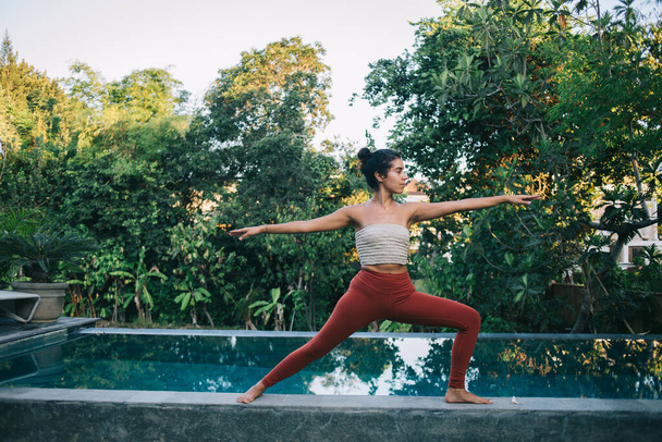 Experienced sportswoman with perfect body shape practising pilates hatha yoga near porch pool doing Virabhadrasana during leisure, good looking female enjoying morning workout for getting inspiration - Fotó, kép
