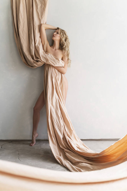 A beautiful blonde curly leggy big breasted naked girl sensually poses at the wall, covering nudity of her excellent body with a large piece of beige cloth. Copy space. Artistic design. - Foto, Imagen