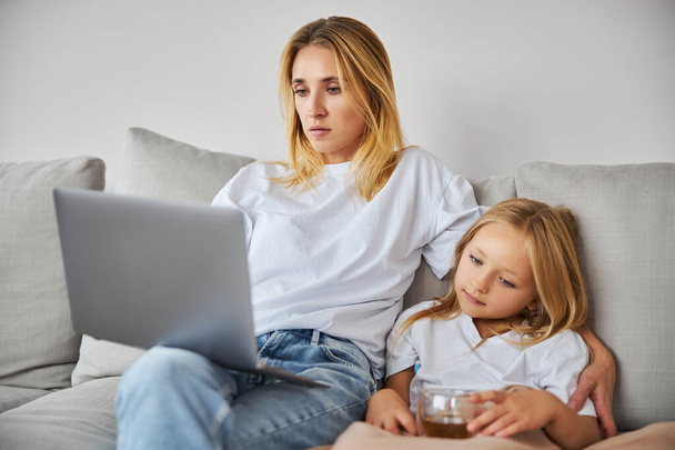 Ernsthaft blonde kaukasische Geschäftsfrau arbeitet am Laptop, während sie mit einem kleinen Mädchen auf dem Sofa sitzt - Foto, Bild