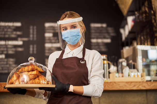 Красивая официантка несет поднос с выпечкой в кафе во время пандемии - Фото, изображение
