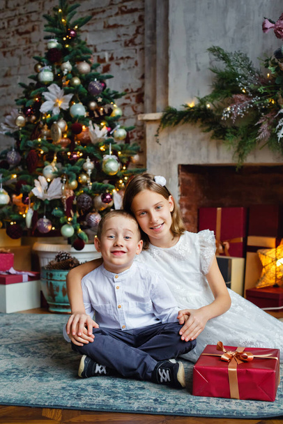 Brother and sister smartly dressed with a gift near the fireplace and Christmas tree with gifts. - Zdjęcie, obraz