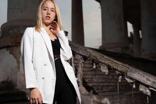 a young blonde eighteen, generation Y, in a white jacket and black pants, standing on the steps of a vintage building. Fashion portrait Fashion model in white jacket on stairs on street. She wears high heels, looking down - Photo, Image