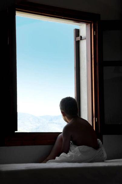 Pieni poika sängyllä katselee ulos ikkunasta, aurinkoisena aamuna lähellä ikkunaa. - Valokuva, kuva