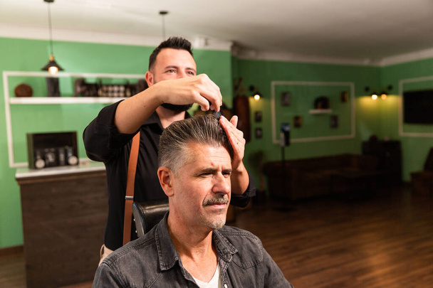 peluquero con camisa y tirantes peine con cepillo y manos al hombre con pelo gris y perilla en una barbería tanto caucásica - Foto, Imagen
