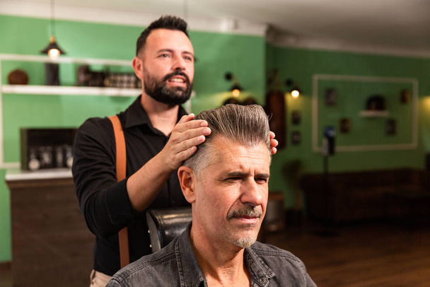peluquero con camisa y tirantes peine con cepillo y manos al hombre con pelo gris y perilla en una barbería tanto caucásica - Foto, Imagen