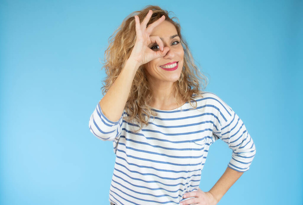 Divertente divertente donna riccia in camicia a righe mostrando gesto ok vicino al suo occhio - Foto, immagini