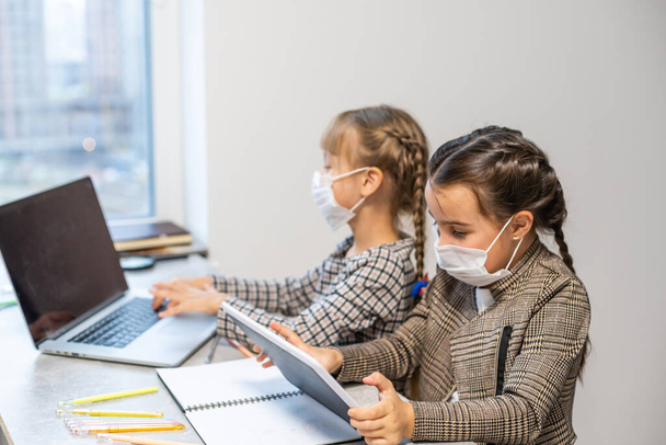 Enseignement à distance. Les filles de l'école à l'ordinateur portable portant masque étudiant assis en ligne à la table à la maison - Photo, image