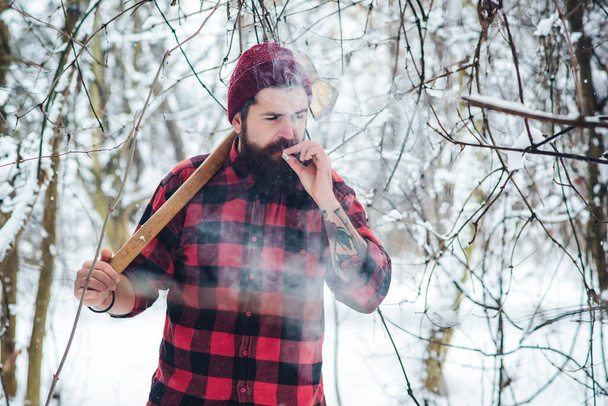 Een man met baard en sigaret. Lumberjack in de winter besneeuwd bos. Een bebaarde hipster met bijl. Serieuze houthakker op winterdag buiten. Houthakker levensstijl. Knappe man die in het bos werkt. - Foto, afbeelding
