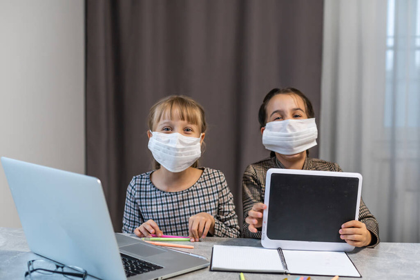 Afstandsonderwijs. School meisjes op laptop dragen masker Studeren Online zitten aan de tafel thuis - Foto, afbeelding