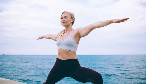 Caucasian female yogi practicing Virabhadrasana during morning workout at fresh ocean air, experienced slim woman in tracksuit doing warrior pose two enjoying healthy lifestyle for body harmony - Foto, imagen