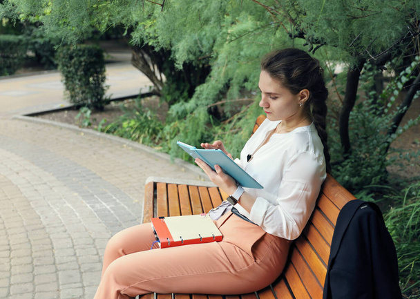 Νεαρή όμορφη μαθήτρια διαβάζει ήρεμα στο τάμπλετ, ενώ κάθεται στο παγκάκι μόνη της. - Φωτογραφία, εικόνα