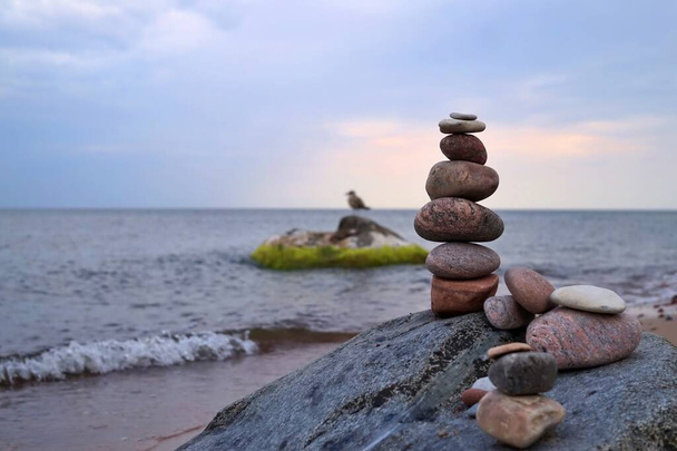 Groot uitgebalanceerd steenmannetje van gestapelde stenen of rotsen op een strand bovenop een rots met uitzicht op de oceaan en zachte golven bij zonsondergang - Foto, afbeelding