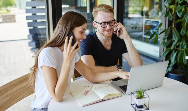 Successful hipster students using smartphone devices for call to customer service and discuss wireless connection, happy male and female talking via cellphones while watching online webinar on laptop - 写真・画像