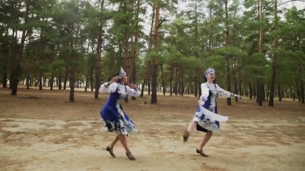 Dos hermosas mujeres en traje popular tradicional ruso azul con pañuelo están bailando en el bosque - Imágenes, Vídeo