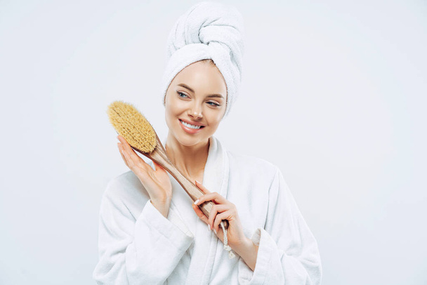 Beautiful glad woman with healthy skin and toothy smile holds wooden massage brush for body, wears bath robe and wrapped soft towel on head, stands against white background. Beauty, care concept - Photo, Image