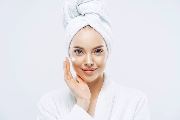 Young confident Caucasian woman applies face cream, enjoys new anti wrinkle cosmetic product, prevents sign of skin aging, wears minimal makeup, dressed in bath robe, isolated on white wall. - Photo, Image