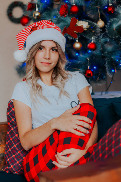 Young beautiful woman in Santa hat sitting on sofa in front of Christmas tree  - Фото, зображення