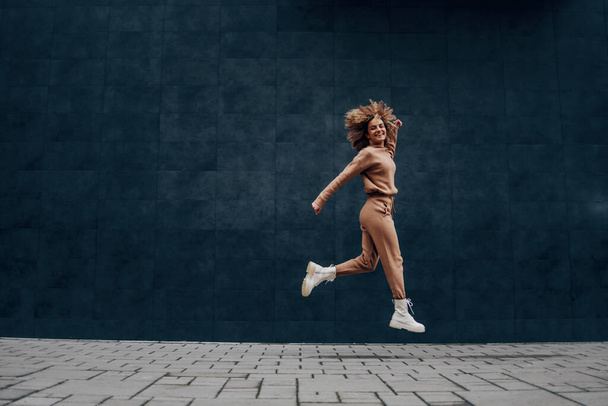 Młoda modna uśmiechnięta kobieta z kręconymi włosami skacząca i biegająca na świeżym powietrzu. - Zdjęcie, obraz