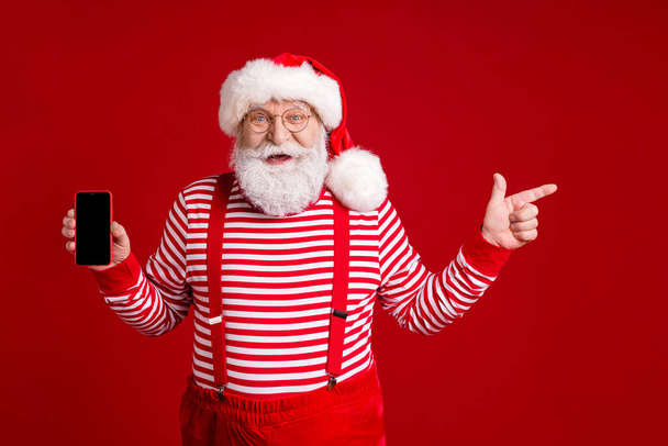 Portrait of his he nice handsome glad cheerful bearded Santa holding in hands device demonstrating advert copy space sale like follow isolated bright vivid shine vibrant red color background - Foto, immagini