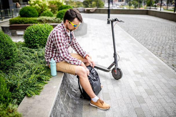 Young caucasian man opens a black roll top backpack while sitting on a bench near an electric scooter in a city park. Courier, delivery service concept. Male with rucksack looks into an open backpack. - 写真・画像